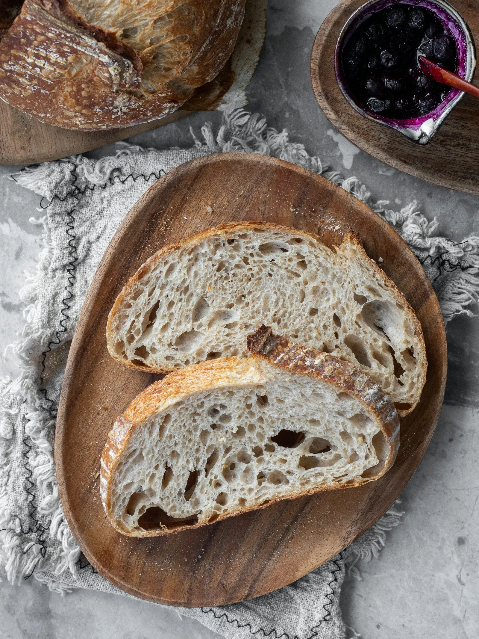 Open-crumb sourdough slices on a round wooden board beside a loaf and jam.
