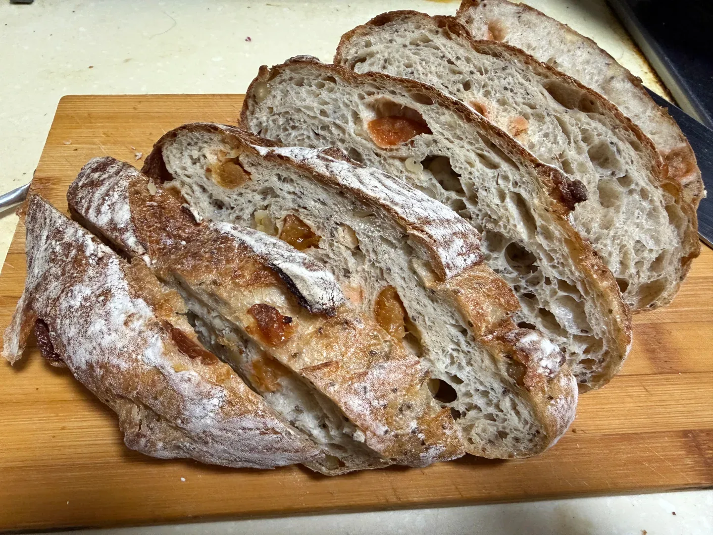 Thick slices of a flour-dusted loaf with golden pieces and dark flecks arranged on a wooden board.