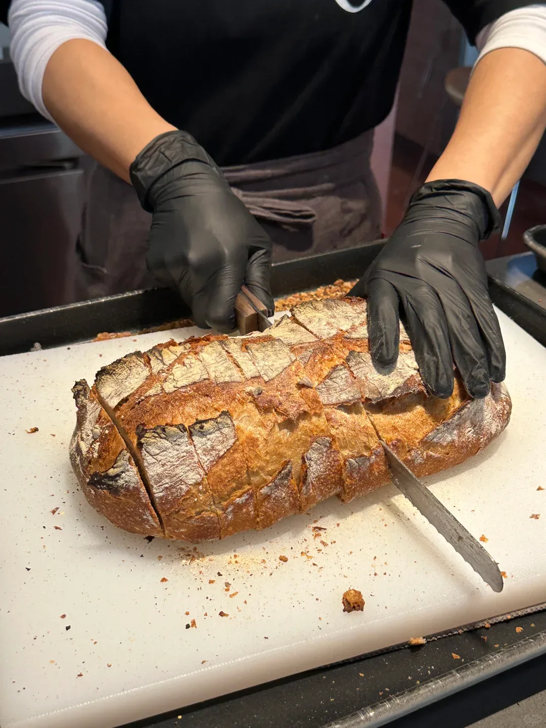 Gloved hands cutting a large golden-crusted loaf into thick slices on a cutting board.